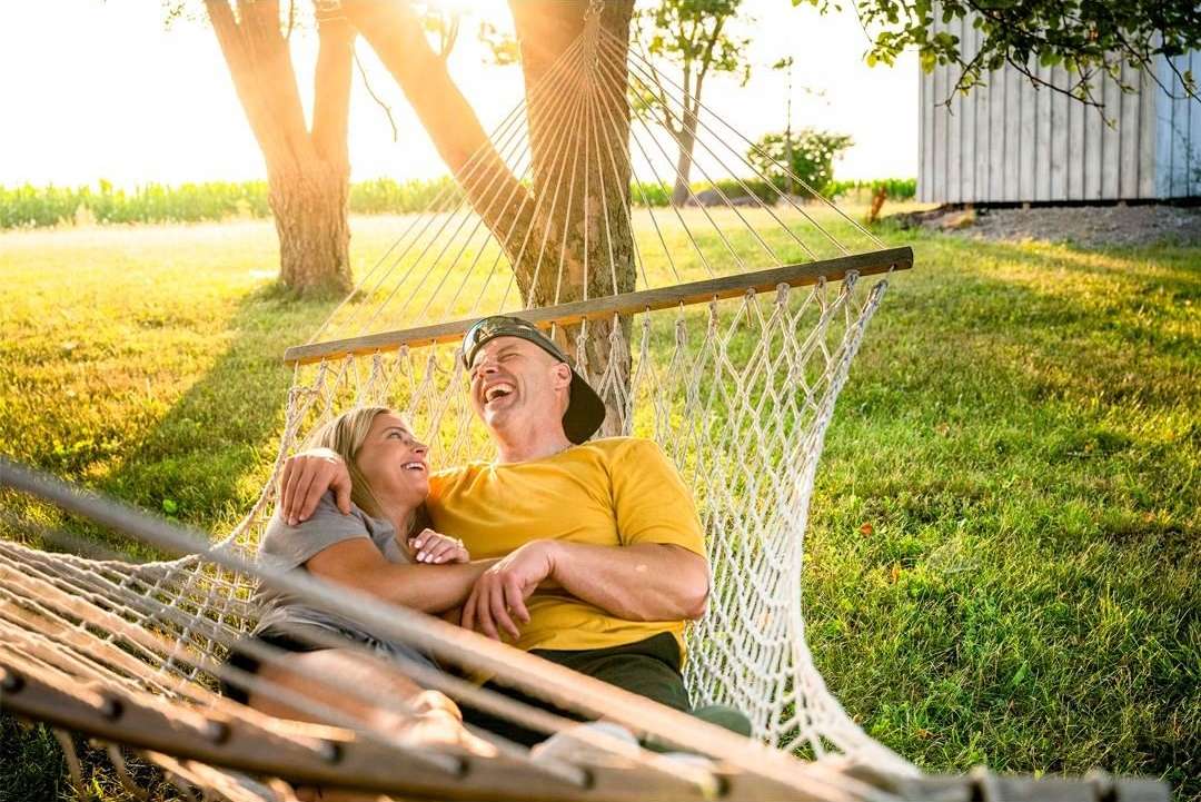 A happy couple relaxes in a hammock outdoors on a sunny day. The woman looks up at the man as he throws his head back laughing.
