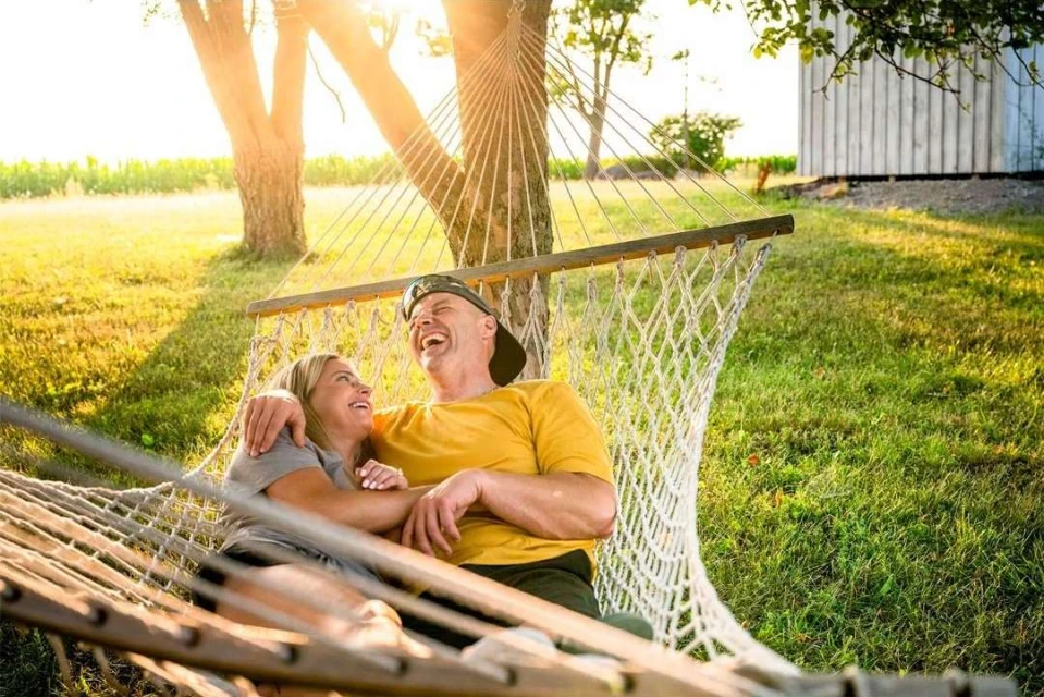 A happy couple relaxes in a hammock outdoors on a sunny day. The woman looks up at the man as he throws his head back laughing.
