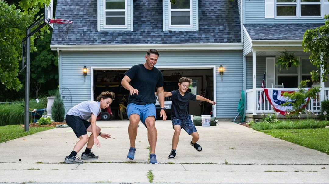Father and two sons playing with a ball in their driveway in front of a two-story light blue house on a bright day outdoors.