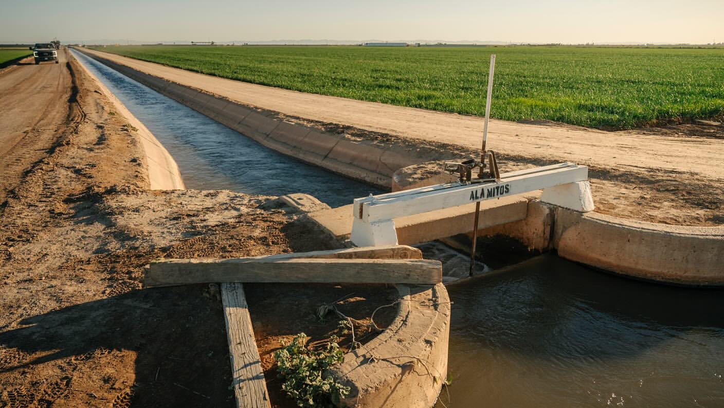 Water canal with concrete barriers and control gate running through agricultural fields under a clear sky, promoting irrigation.