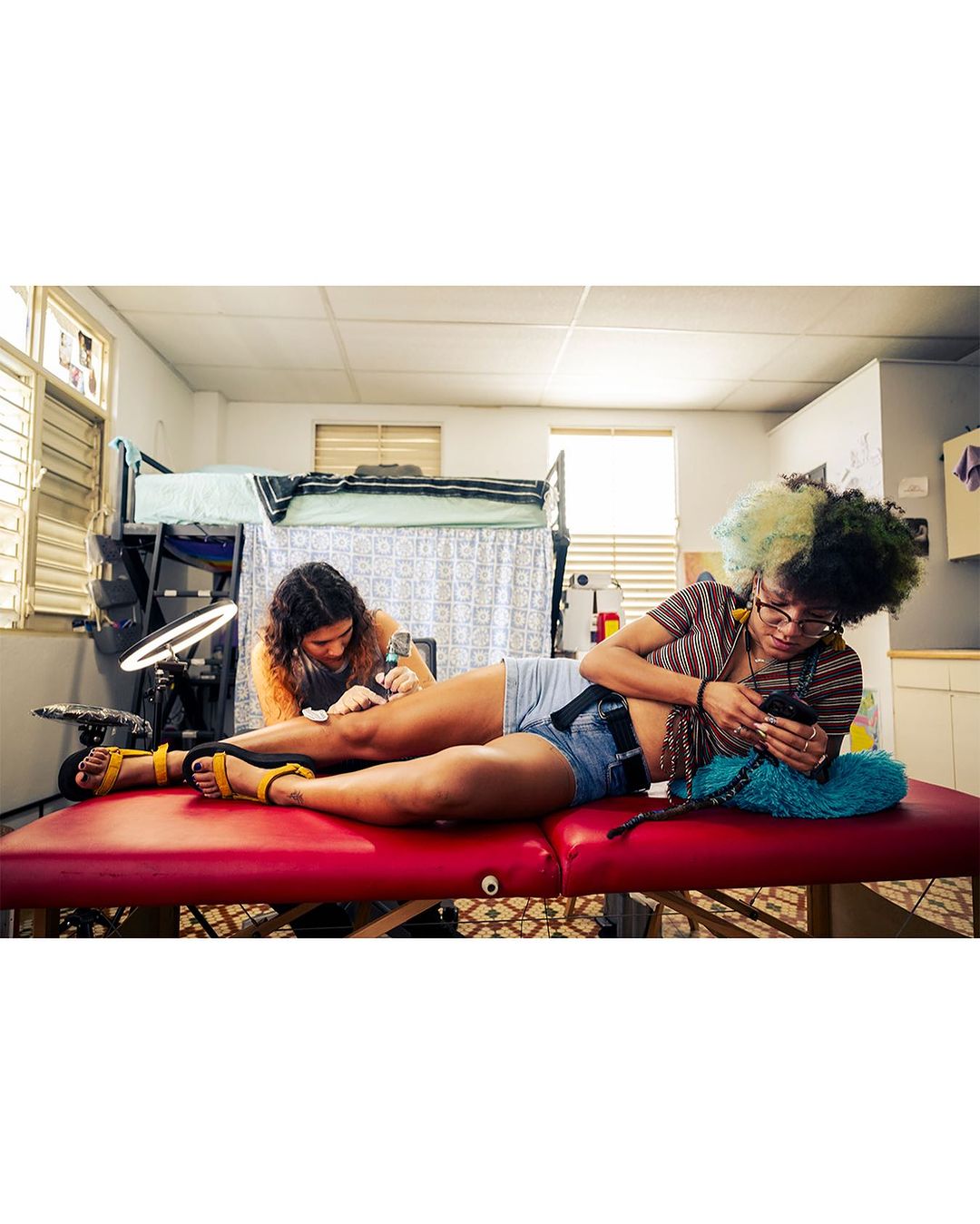 Two women indoors. One woman tattoos the leg of another woman lying on a red examination table while looking at a mobile phone.
