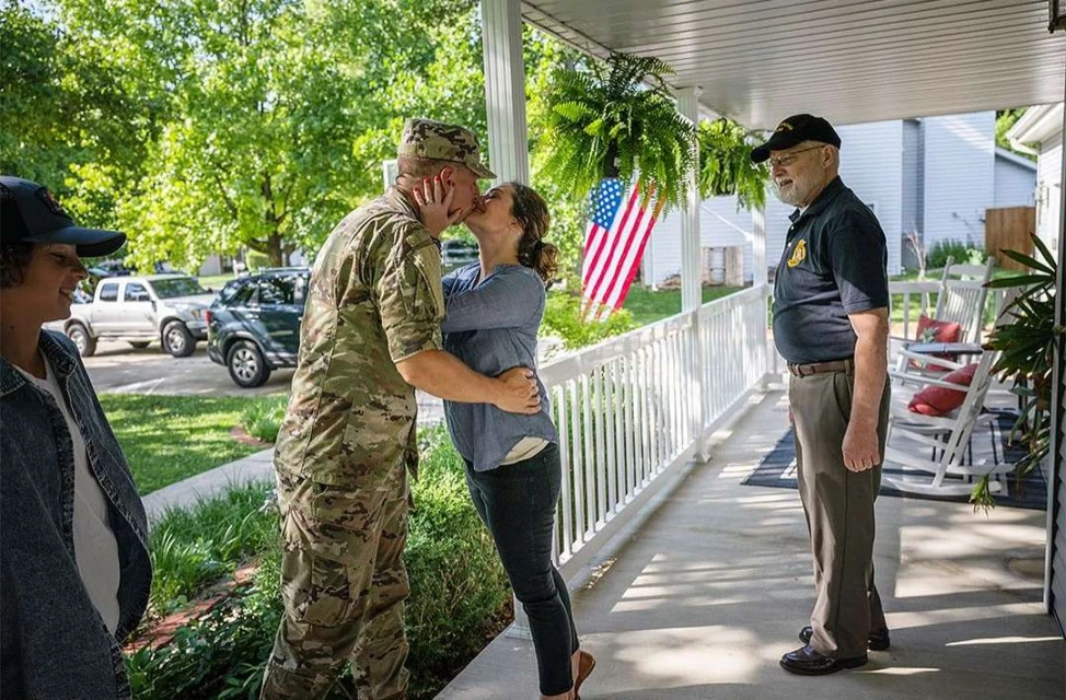 A military member is kissing a woman on a porch with an American flag and a man and woman nearby, standing outside on a sunny day.