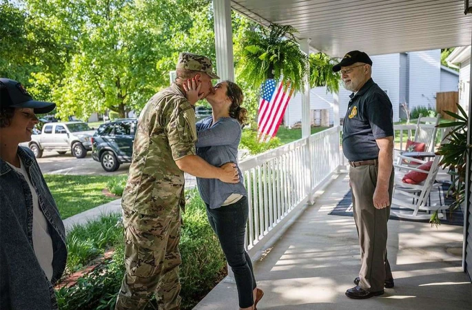 A military member is kissing a woman on a porch with an American flag and a man and woman nearby, standing outside on a sunny day.