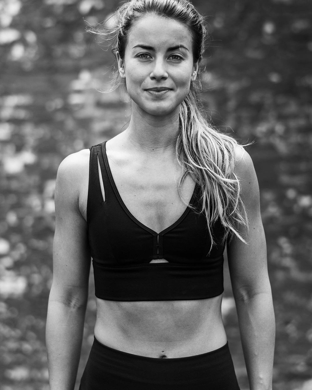 Black and white portrait of a woman in a black sports bra with a ponytail, captured in a medium shot with a blurred background.