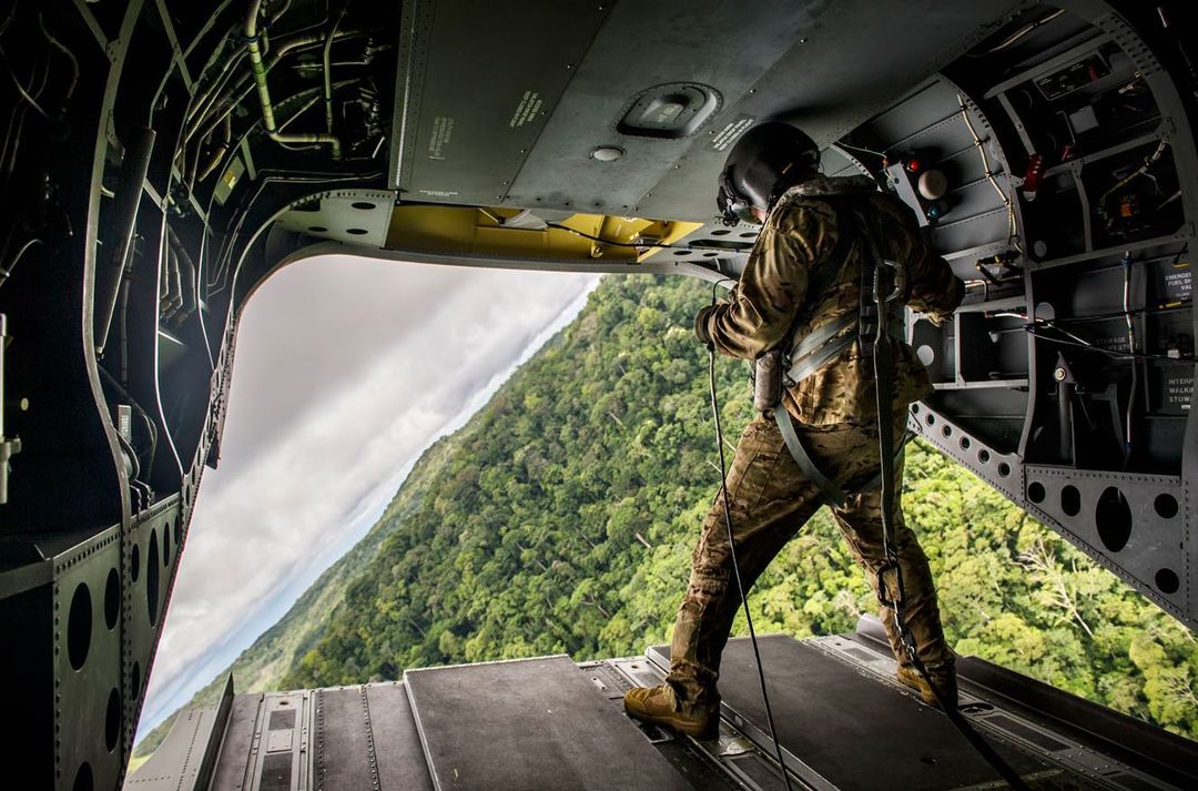 A man in military gear stands on a helicopter ramp with mountains and forest in the background, preparing for a skydive.