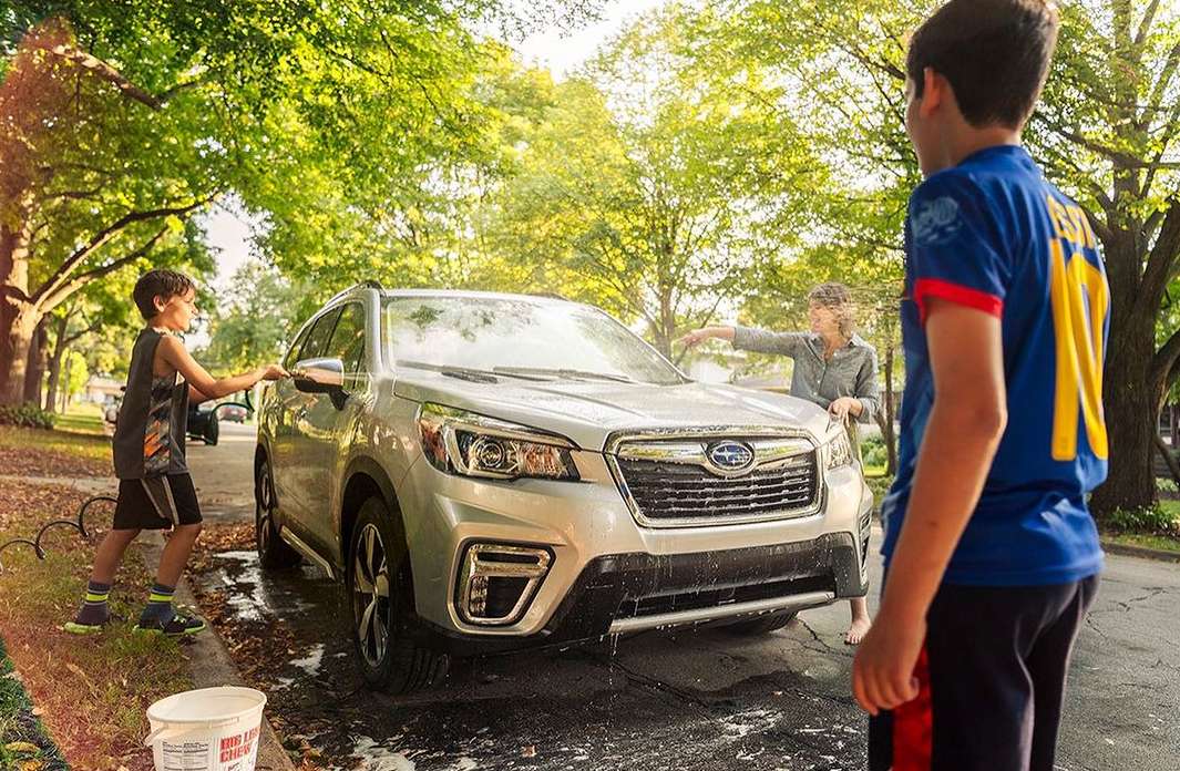 Silver Subaru being washed on a sunny residential street by a mother and two children, with trees and houses in the background.