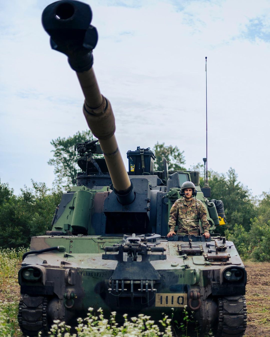 Camouflage military tank in rural setting with soldier in the driver's seat in daylight against a green and cloudy sky.