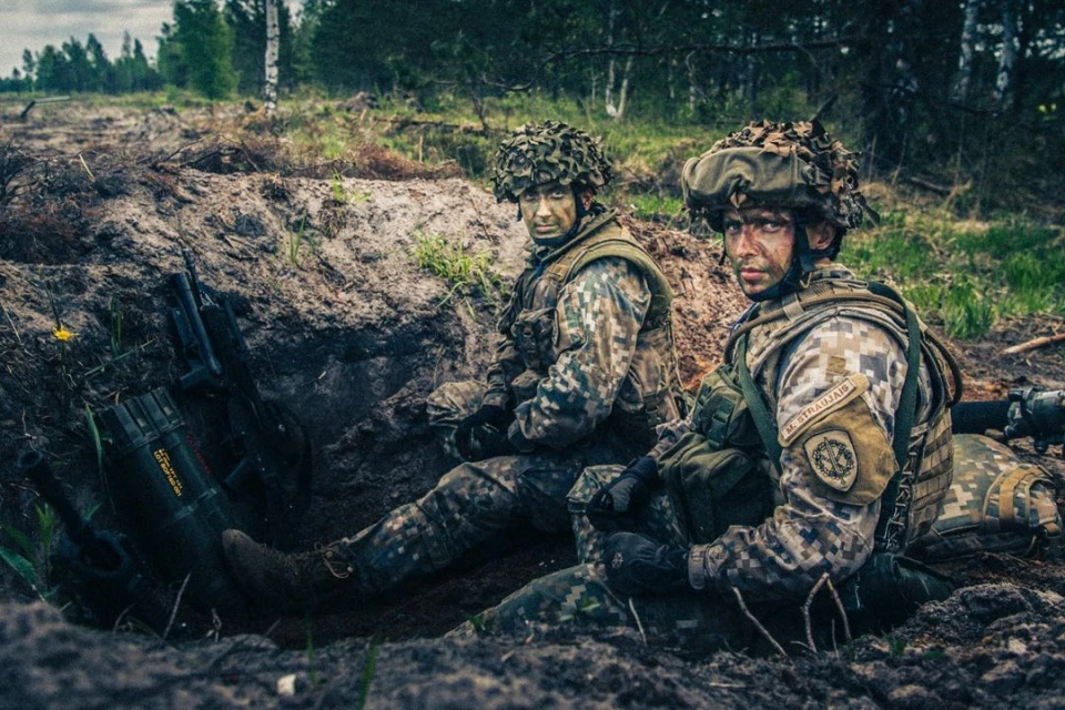 Two soldiers in camouflage uniforms and helmets sit in a trench, surrounded by dirt and vegetation, looking towards the viewer.