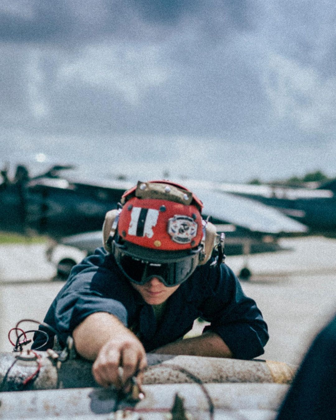 Navy mechanic in helmet and goggles repairs aircraft parts outdoors, with fighter jet visible in the cloudy background.