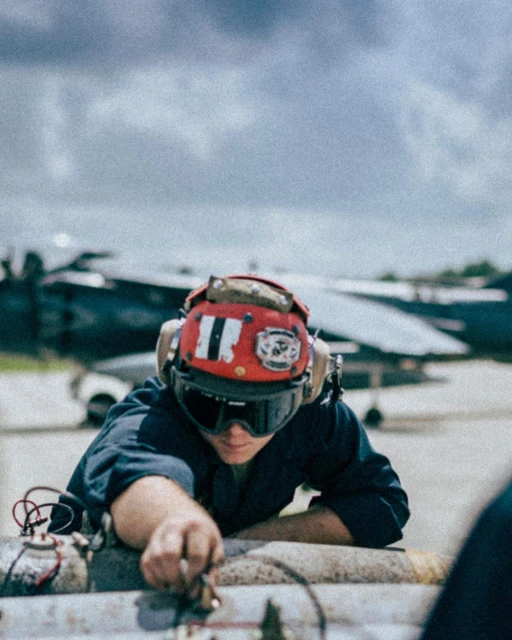 Navy mechanic in helmet and goggles repairs aircraft parts outdoors, with fighter jet visible in the cloudy background.