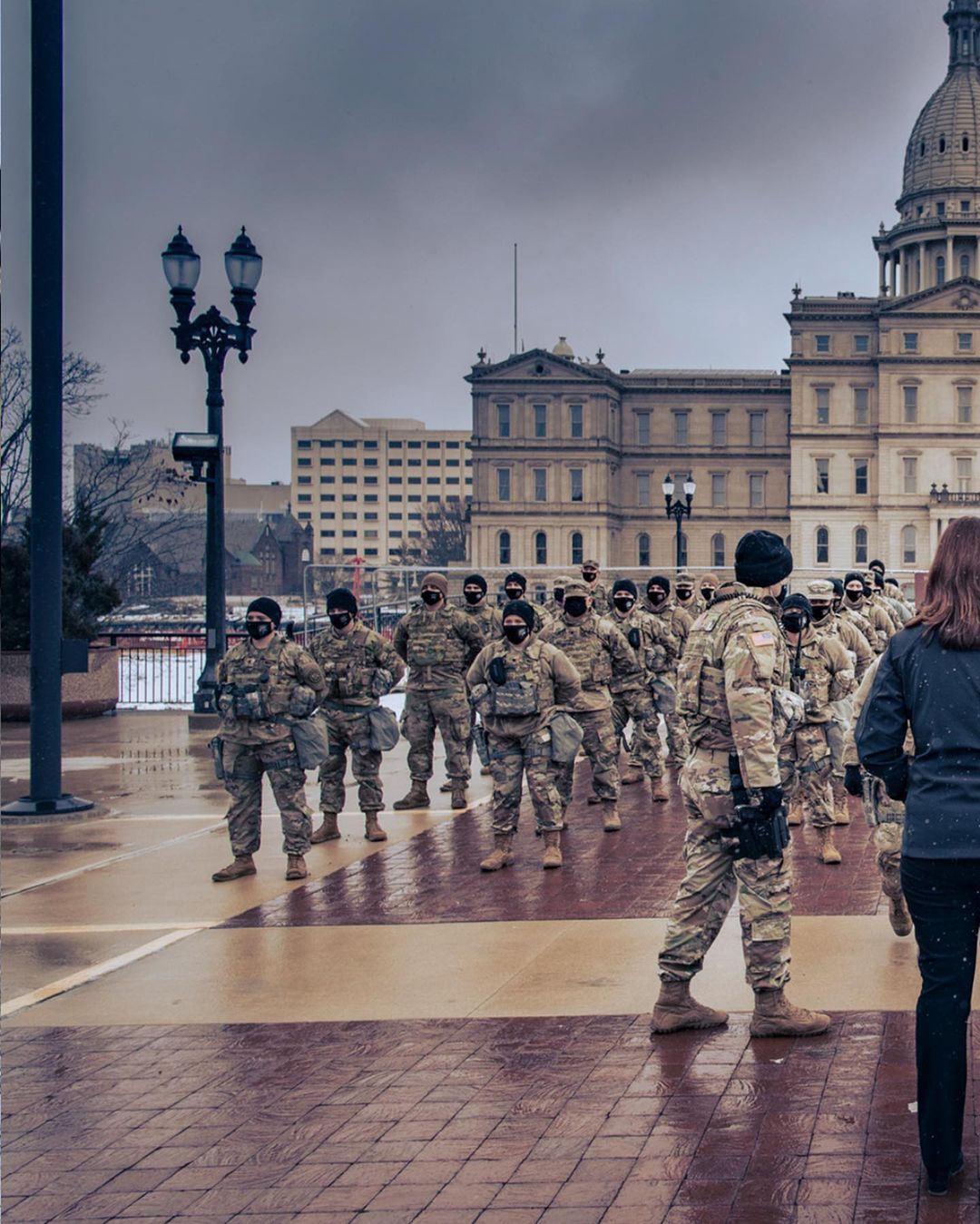 Group of soldiers in camouflage uniforms stand in formation on a brick plaza in front of a capitol building under a cloudy sky.