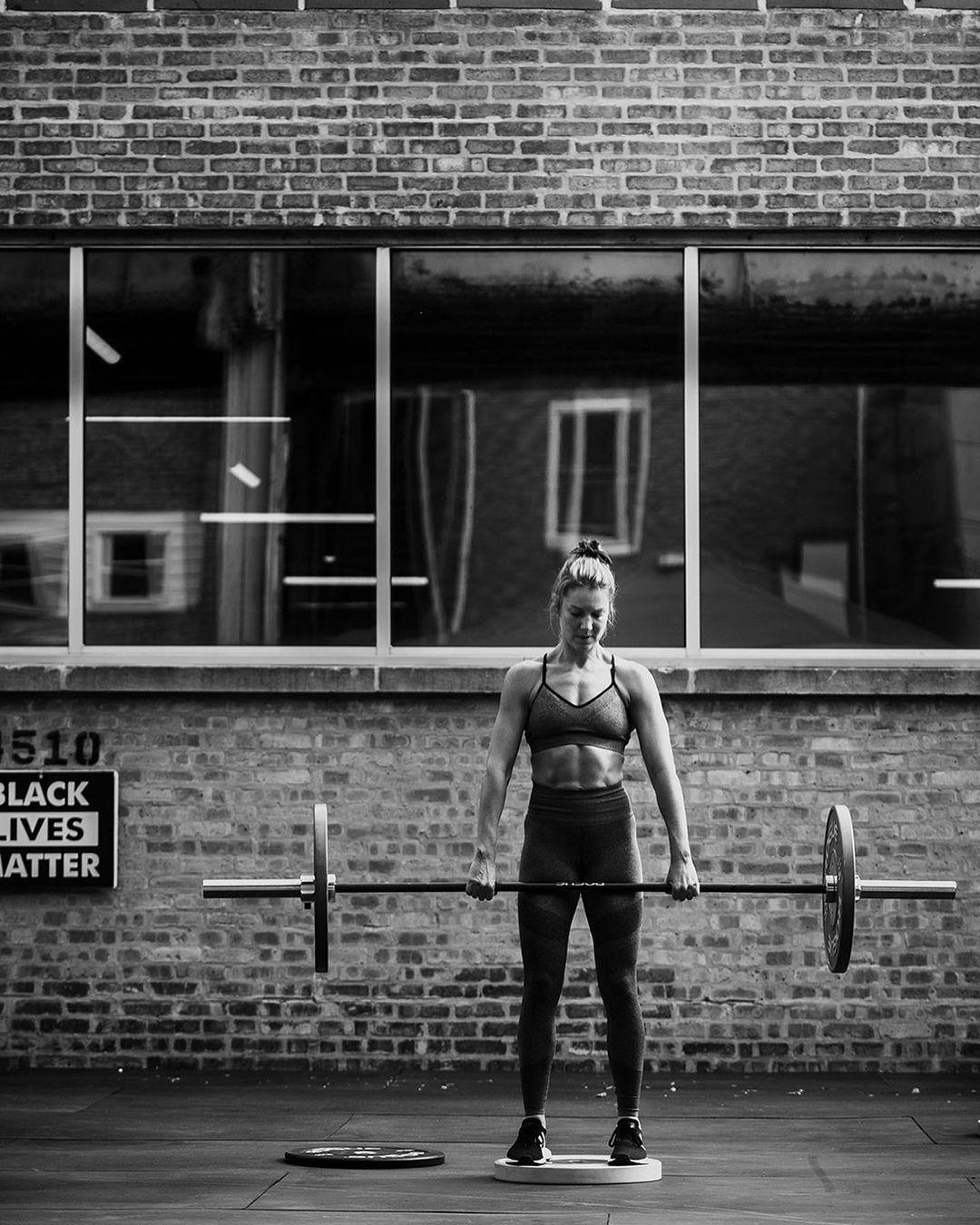 Black and white shot of a strong woman lifting barbell weights in a gym with brick walls and large windows behind.