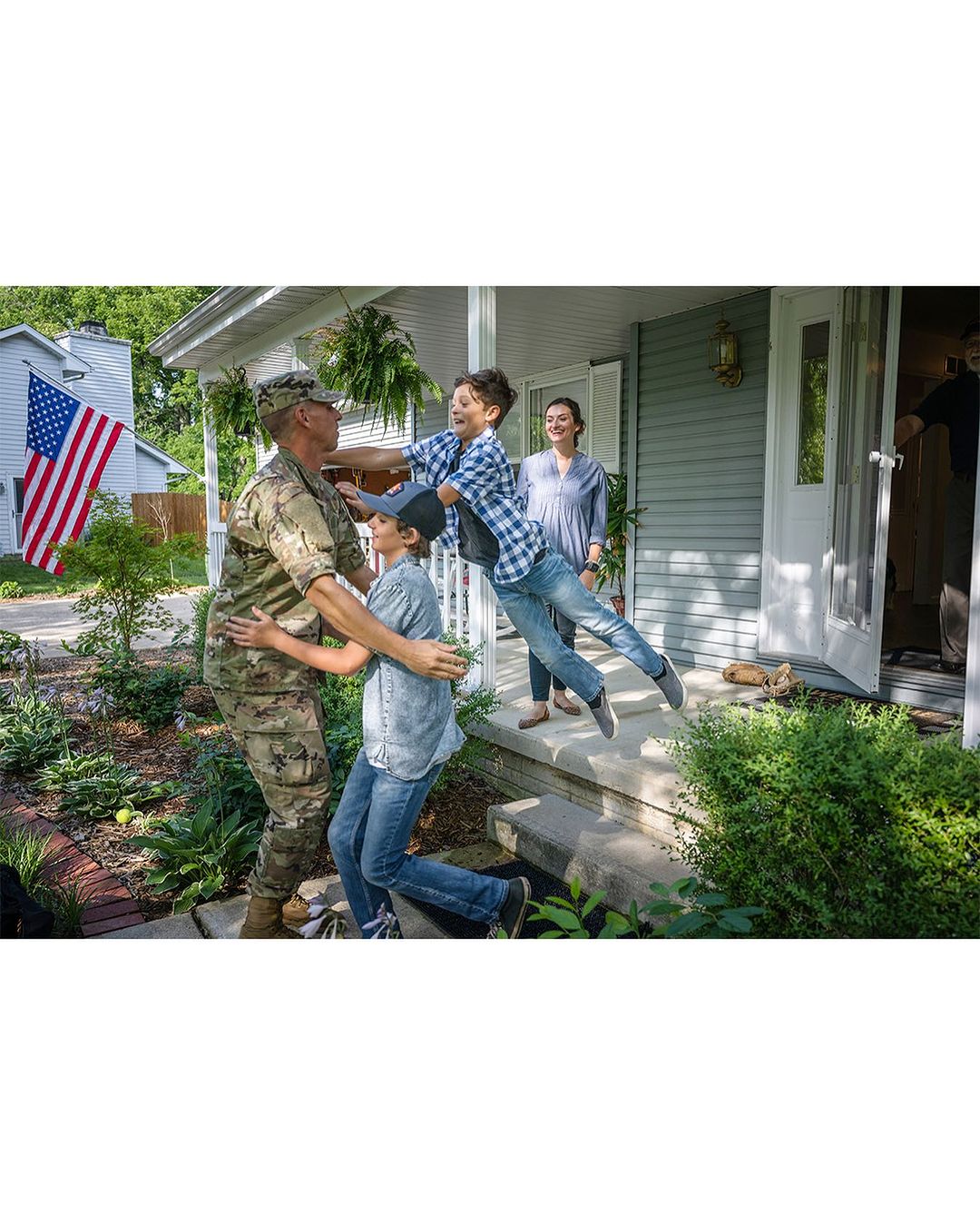 Military personnel embracing two children outside a house, with a woman standing in the background and an American flag on display.