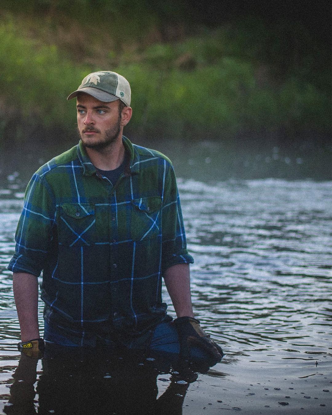 Man standing in a river wearing a flannel shirt, baseball cap, and gloves, surrounded by flowing water and distant green foliage.