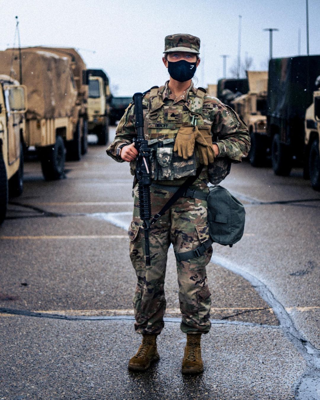 Full shot of military personnel in camouflage uniform with rifle, standing on pavement in an outdoor environment with military vehicles.