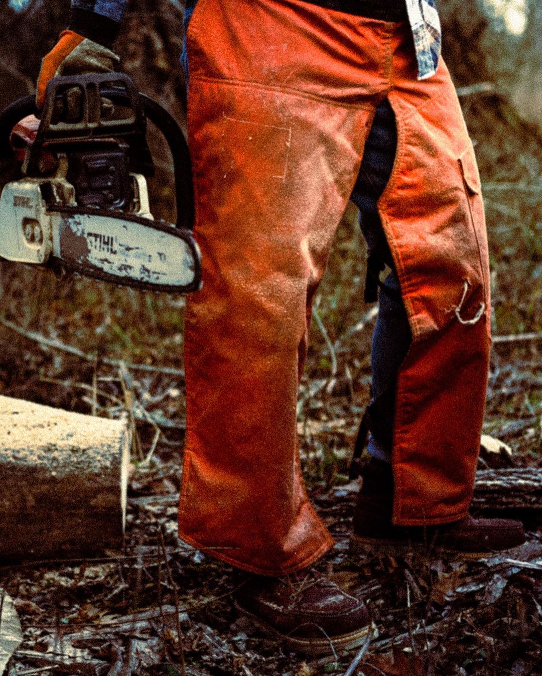 Lumberjack in safety chaps holding a chainsaw near cut logs in a forest environment featuring earth tones and natural lighting.