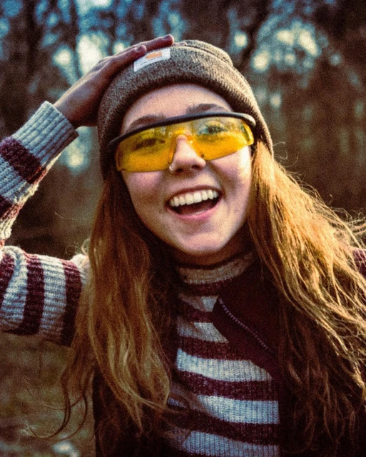 A young woman in a beanie and yellow glasses smiles outdoors, holding her hand on her head, with trees blurred in the background.