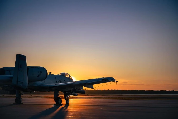 A-10 Thunderbolt II aircraft stands on a runway as the sun rises, silhouetting the plane against the orange and blue sky, creating a dramatic scene.