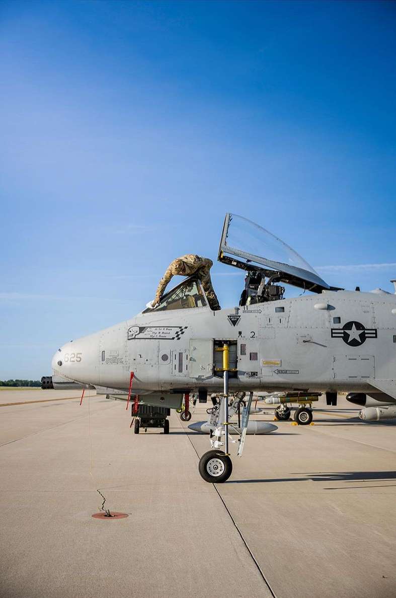 Military personnel performs maintenance on a gray A-10 Thunderbolt II aircraft on an airbase runway with a clear blue sky overhead.