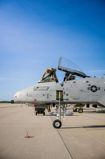 Military personnel performs maintenance on a gray A-10 Thunderbolt II aircraft on an airbase runway with a clear blue sky overhead.