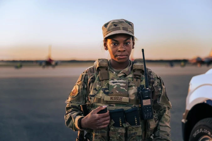 Woman in military uniform stands on airfield at sunset with aircraft and vehicle. The sky is clear with an orange-yellow horizon.