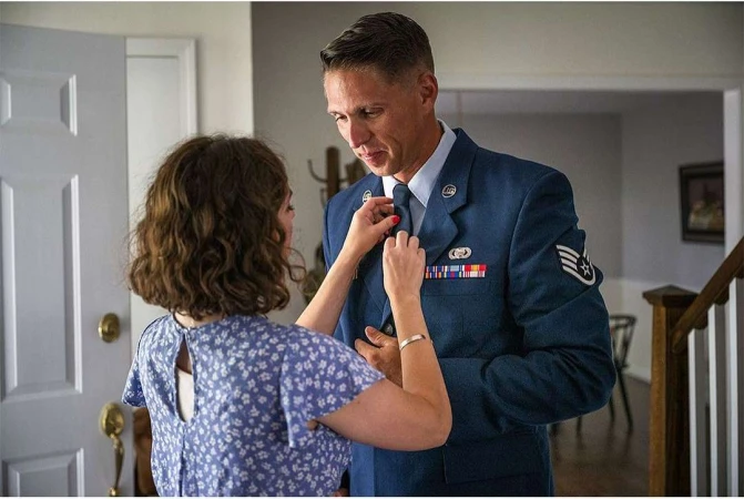 Man in Air Force uniform being helped with his tie by a woman in a floral dress, indoors with a domestic setting and natural lighting.