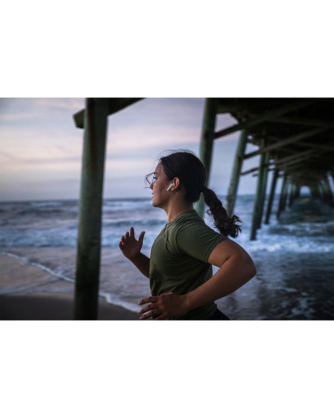 An adult female jogging on a beach near a pier at sunset. She is wearing glasses and earbuds, and has her hair in a ponytail.
