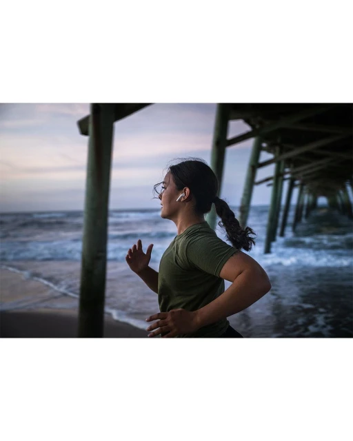 An adult female jogging on a beach near a pier at sunset. She is wearing glasses and earbuds, and has her hair in a ponytail.