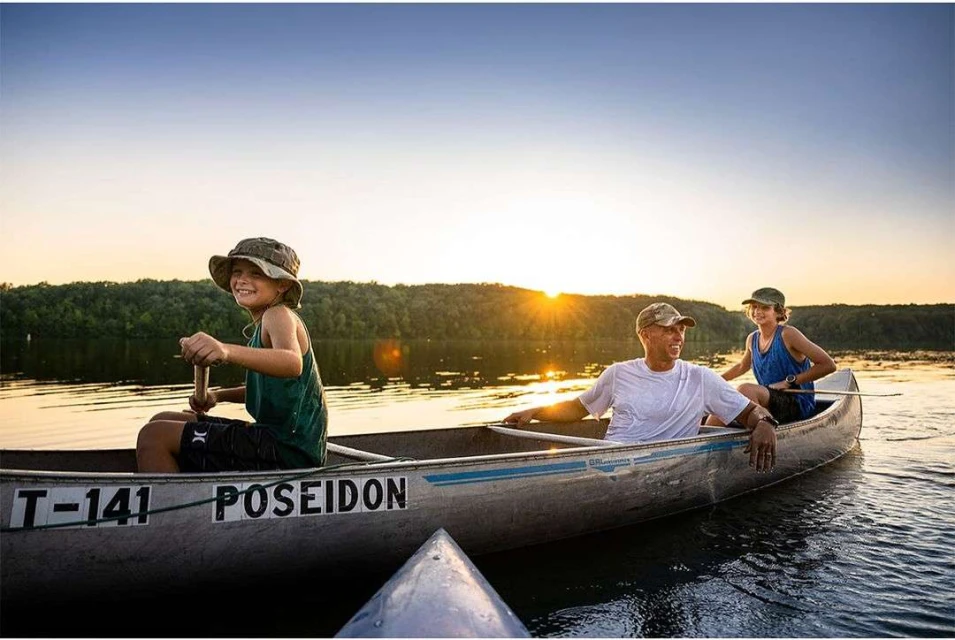 A sunny, outdoor shot depicts a child paddling a canoe, alongside an adult and another child, on a lake with forested shoreline.