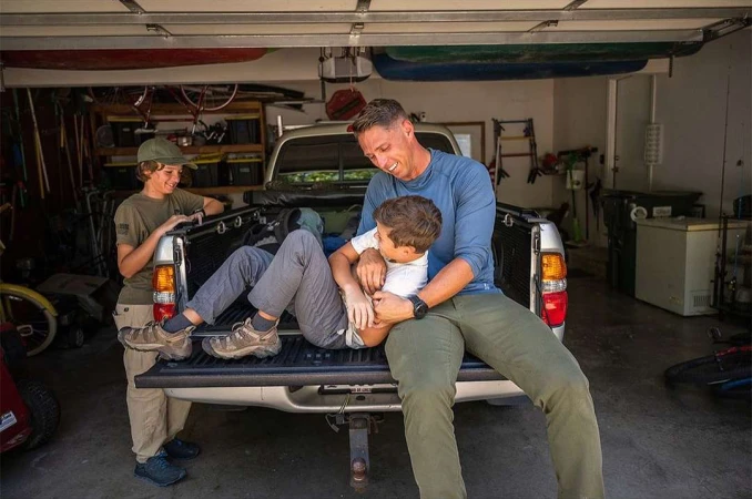 A father and two sons are seated in the back of a parked pickup truck inside a garage, casual clothing, and natural daylight.