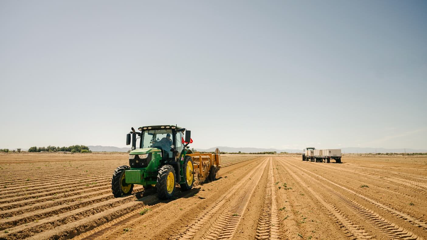 Agricultural scene featuring a green tractor and a trailer in a plowed field under a clear sky, conveying a sense of rural farming activity.