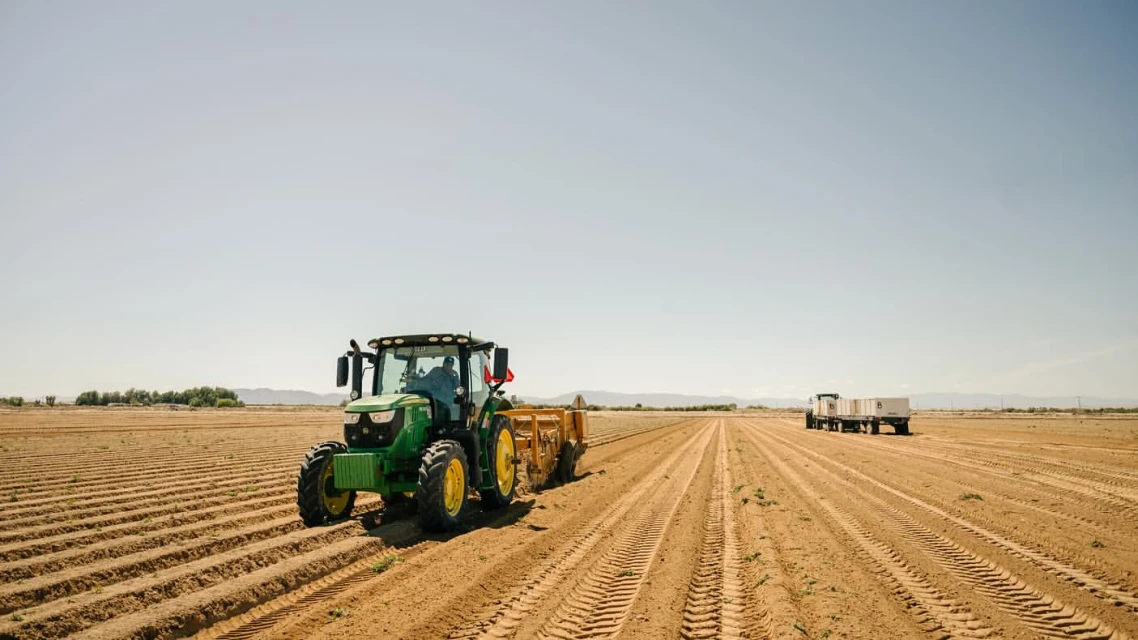 Agricultural scene featuring a green tractor and a trailer in a plowed field under a clear sky, conveying a sense of rural farming activity.