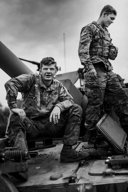Two soldiers in camouflage uniforms are positioned on a military tank against an overcast sky, in a black and white photograph.