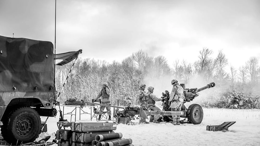 Black and white image of soldiers operating a howitzer in a snowy field with a military vehicle and trees in the background.