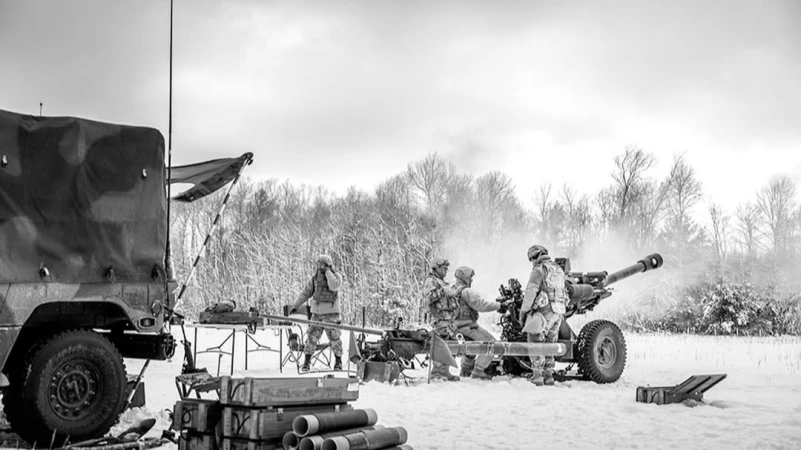 Black and white image of soldiers operating a howitzer in a snowy field with a military vehicle and trees in the background.