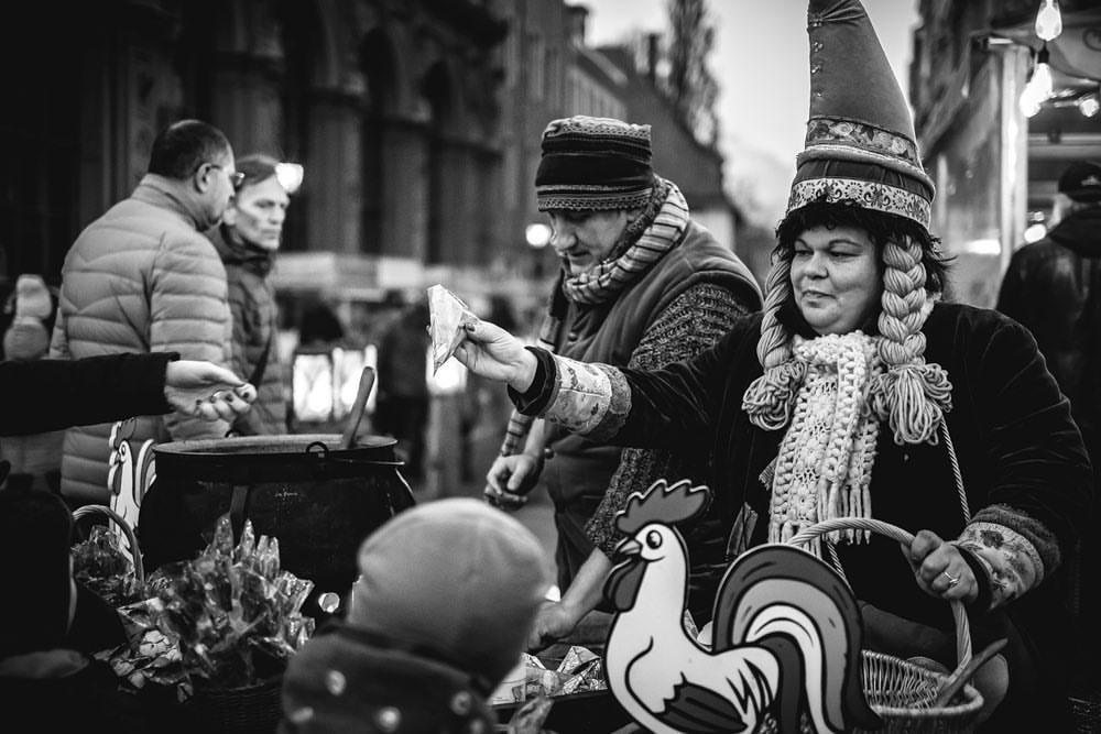 Black and white photo of two people in festive hats at an outdoor market, one holding money and the other holding a basket.