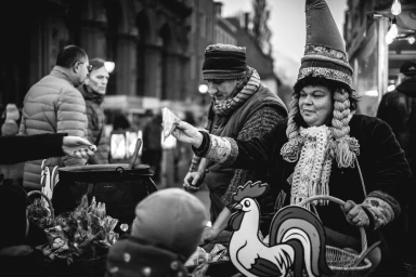 Monochrome street scene of a vendor in a pointed cap handing an item to a customer near a cauldron and rooster cutout
