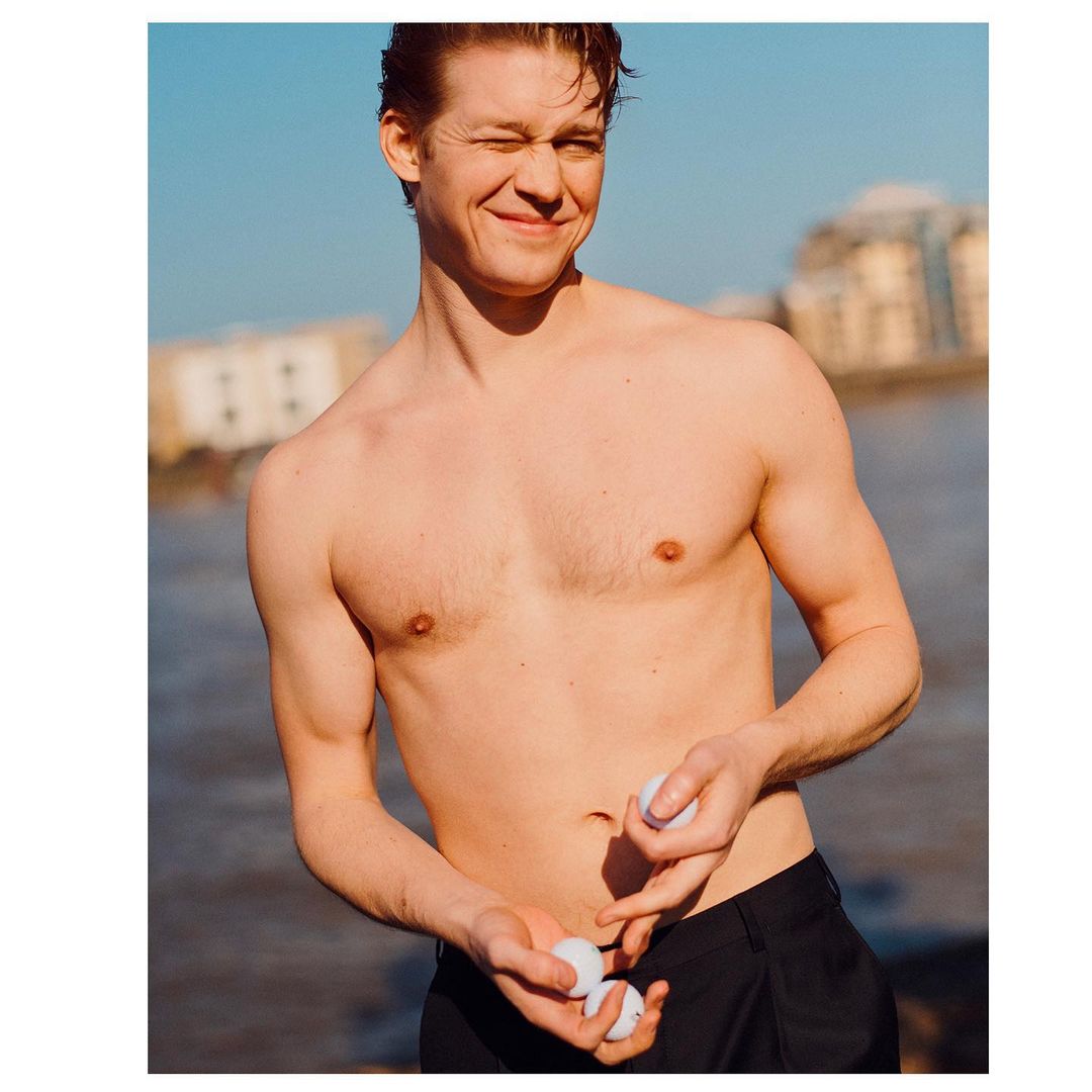 A shirtless, smiling man in dark pants holds white golf balls near a body of water and a building on a sunny day.