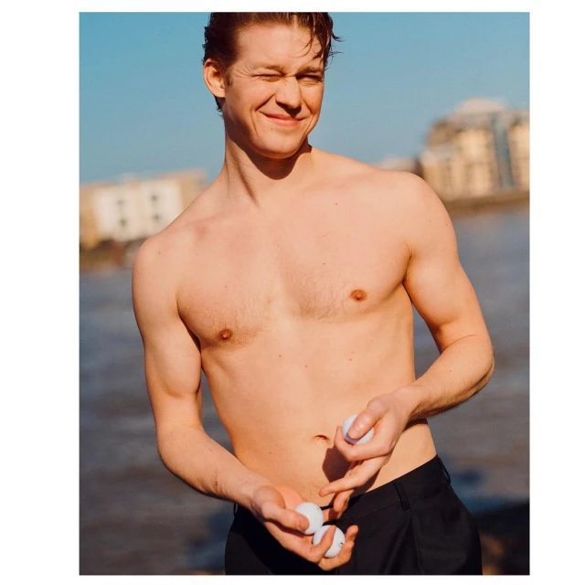 A shirtless, smiling man in dark pants holds white golf balls near a body of water and a building on a sunny day.