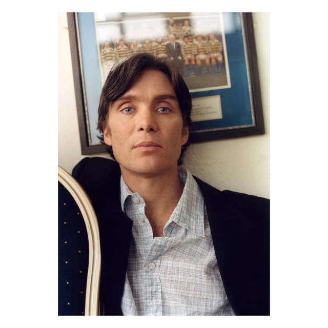 Cillian Murphy portrait indoors, wearing a light-blue patterned button-down shirt and a black blazer, posing against a picture frame on the wall.