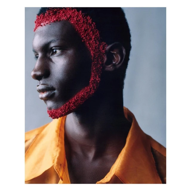 Portrait of an adult Black male model wearing an orange shirt with red textured makeup around his hairline and sideburns.