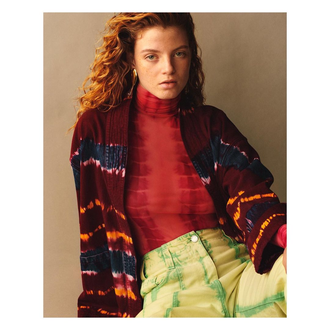 A young adult woman with curly red hair poses indoors wearing a red top, a burgundy jacket with colorful stripes, and chartreuse pants.