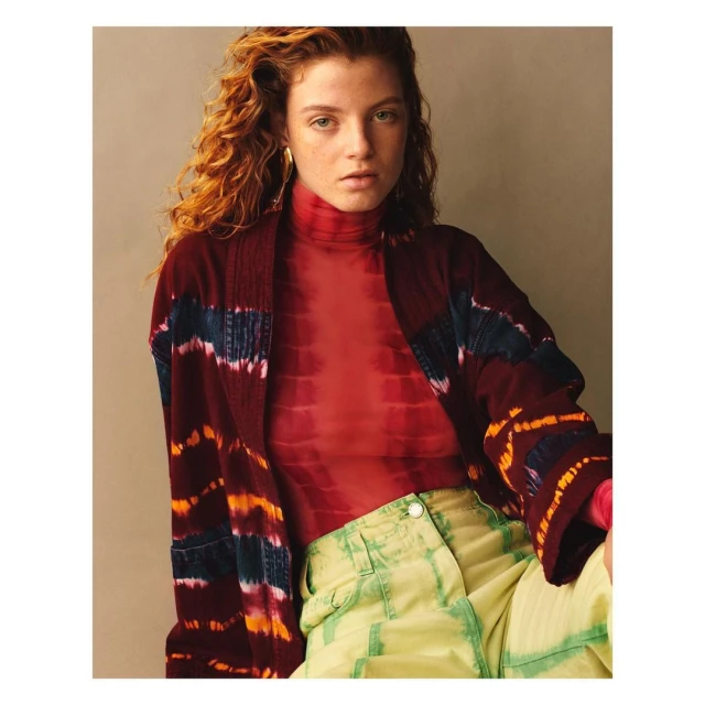 A young adult woman with curly red hair poses indoors wearing a red top, a burgundy jacket with colorful stripes, and chartreuse pants.