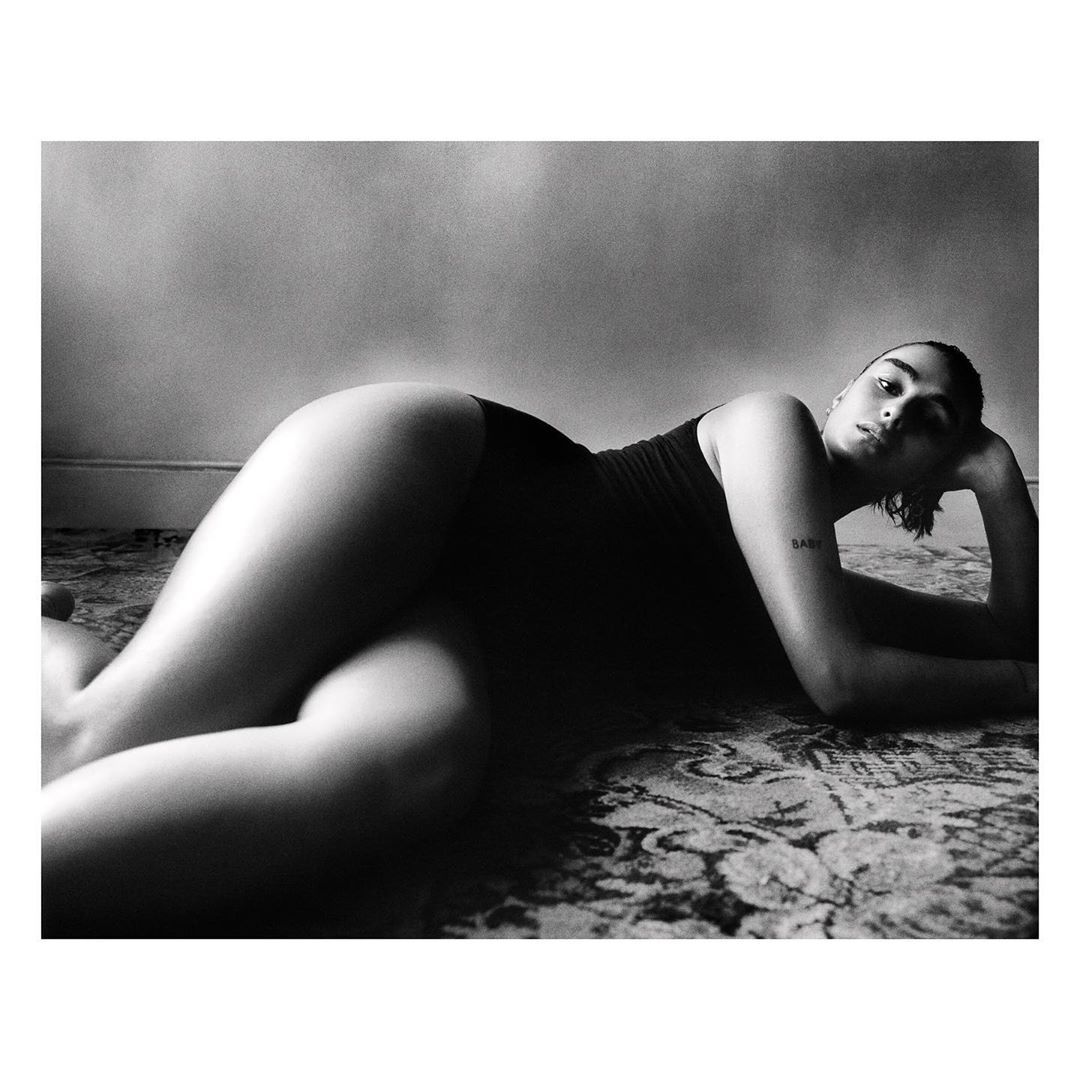 Black-and-white portrait of a woman in a dark bodysuit, lying on a patterned rug, propped up by her elbow, with a neutral facial expression.