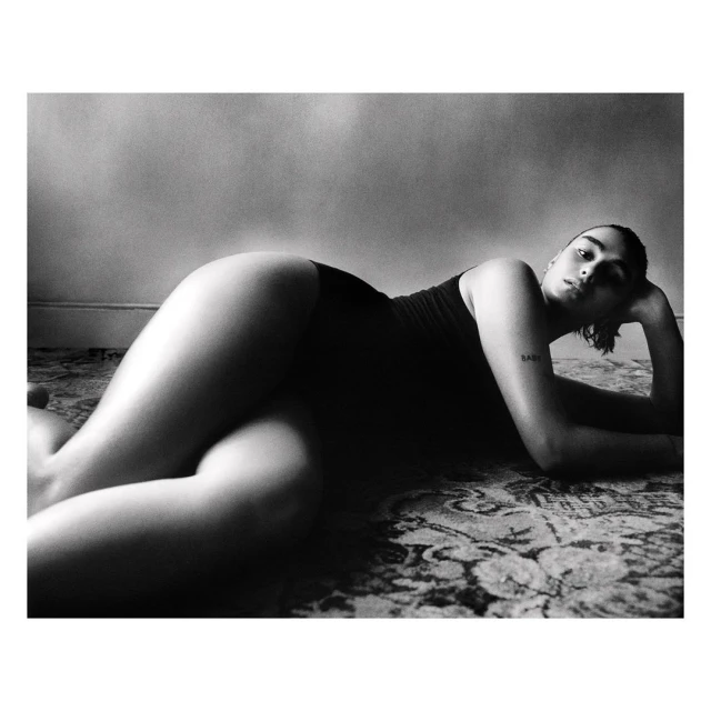 Black-and-white portrait of a woman in a dark bodysuit, lying on a patterned rug, propped up by her elbow, with a neutral facial expression.