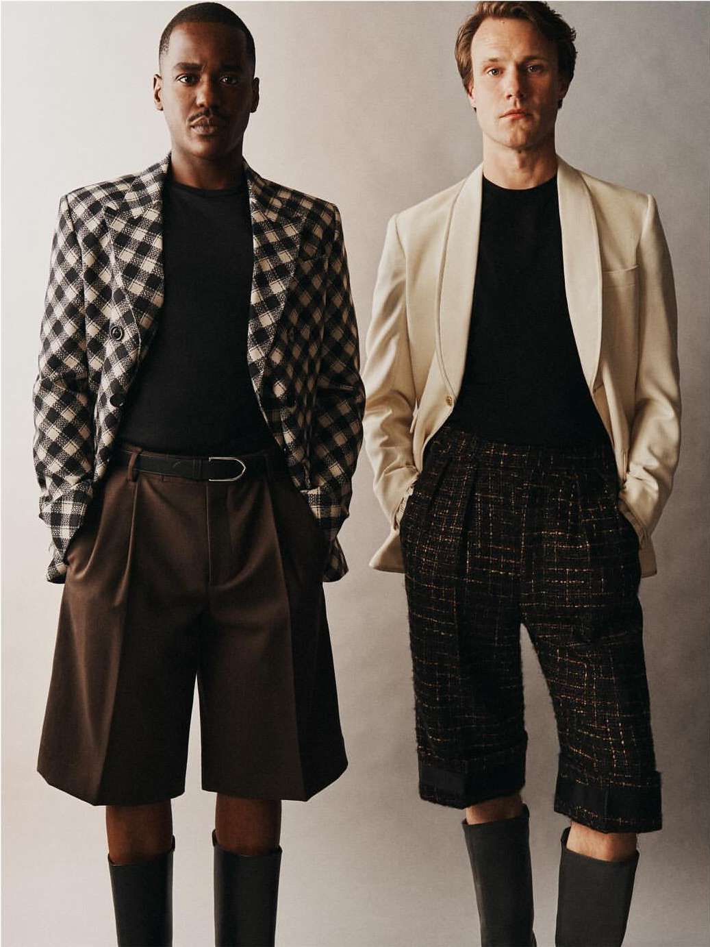 Two men stand side-by-side wearing blazers, black shirts, brown shorts, and boots against a neutral background in a fashion shot.