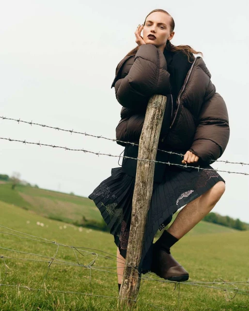 Fashion editorial shot of a woman in a brown puffer jacket leaning against a barbed wire fence in a rural landscape with a grassy background.