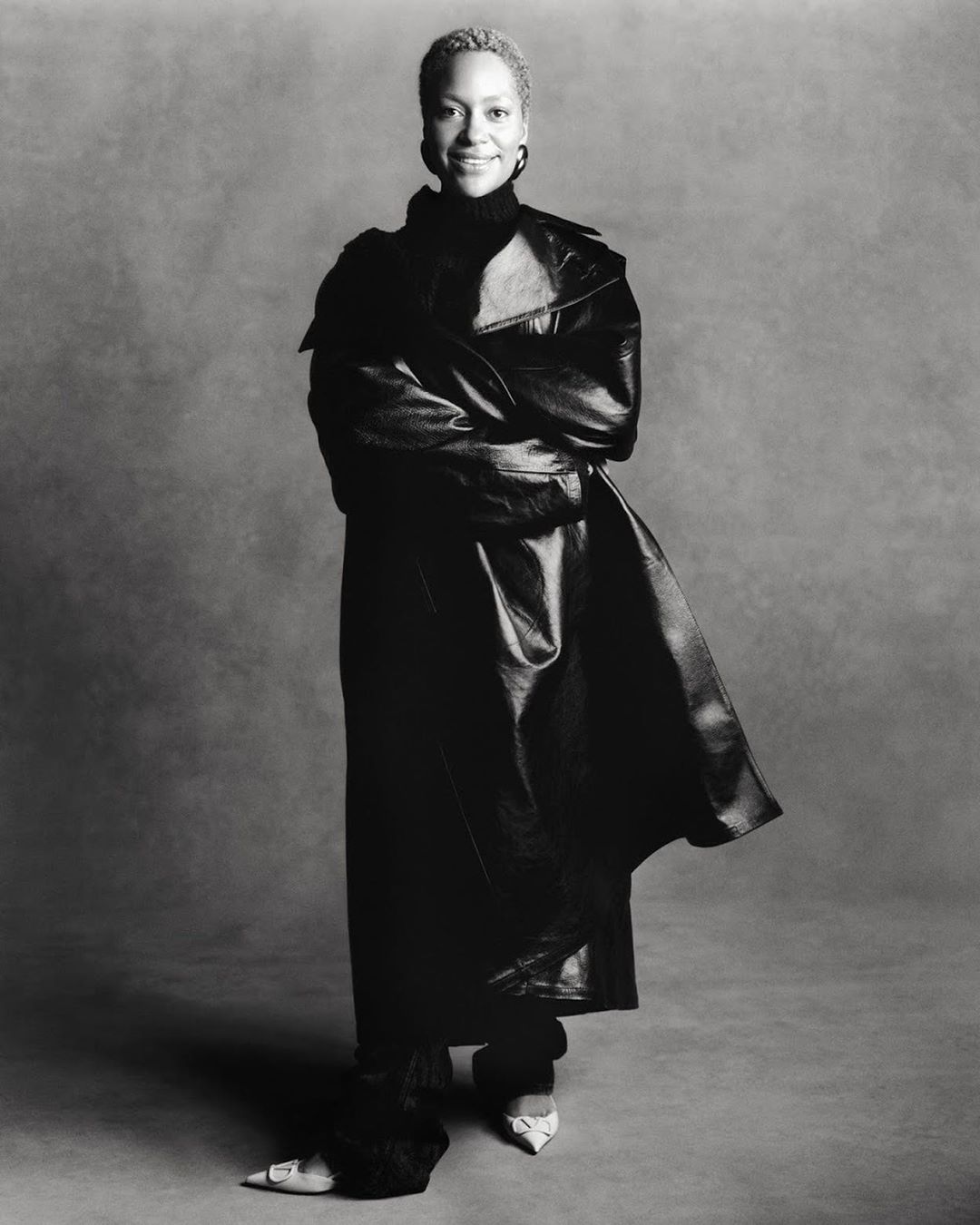 Black and white portrait of a smiling adult woman in a leather coat and dark clothing against a grey backdrop in a studio.