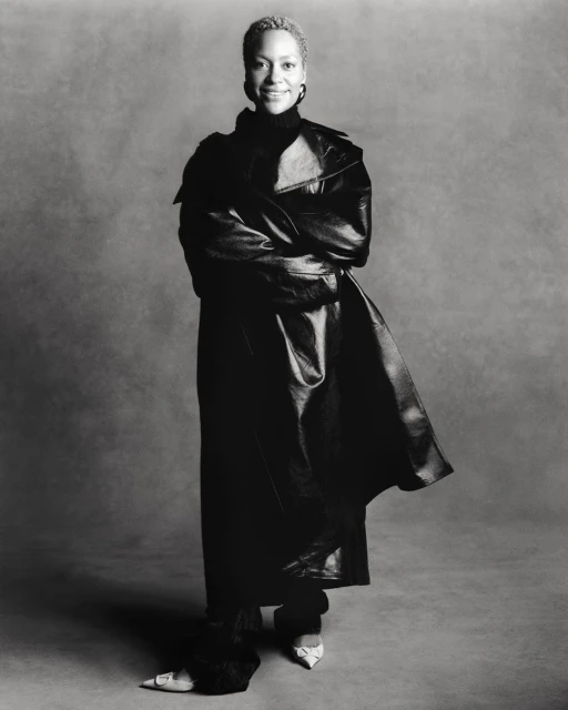 Black and white portrait of a smiling adult woman in a leather coat and dark clothing against a grey backdrop in a studio.