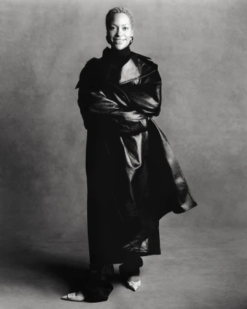 Black and white portrait of a smiling adult woman in a leather coat and dark clothing against a grey backdrop in a studio.